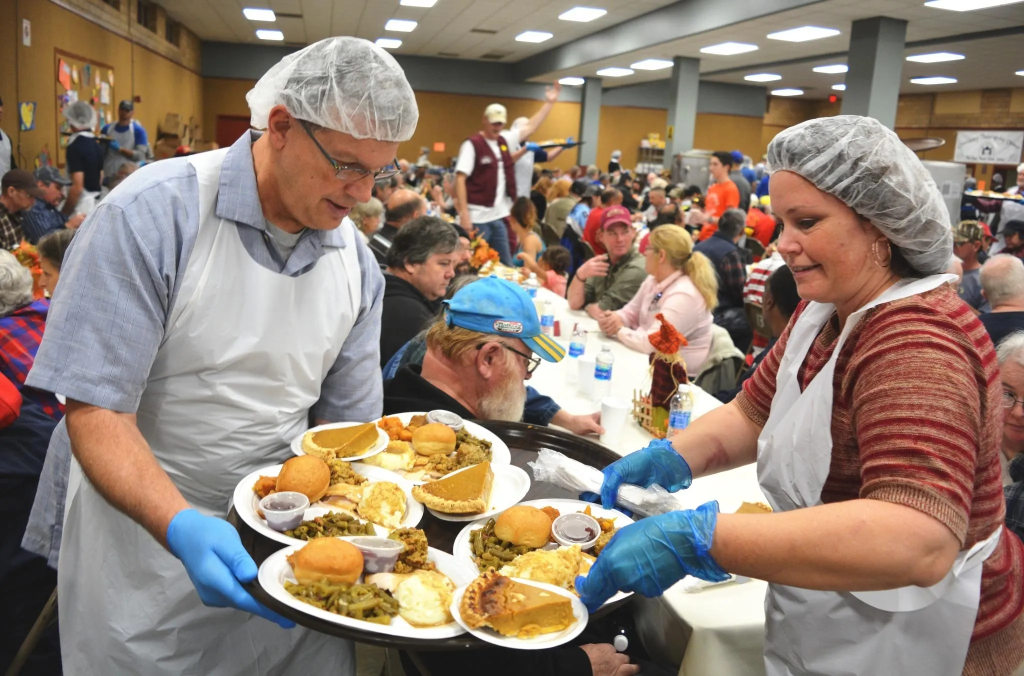 A volunteer in a blue shirt holds a tray of Thanksgiving dinners while a volunteer in a red striped shirt picks up plates.